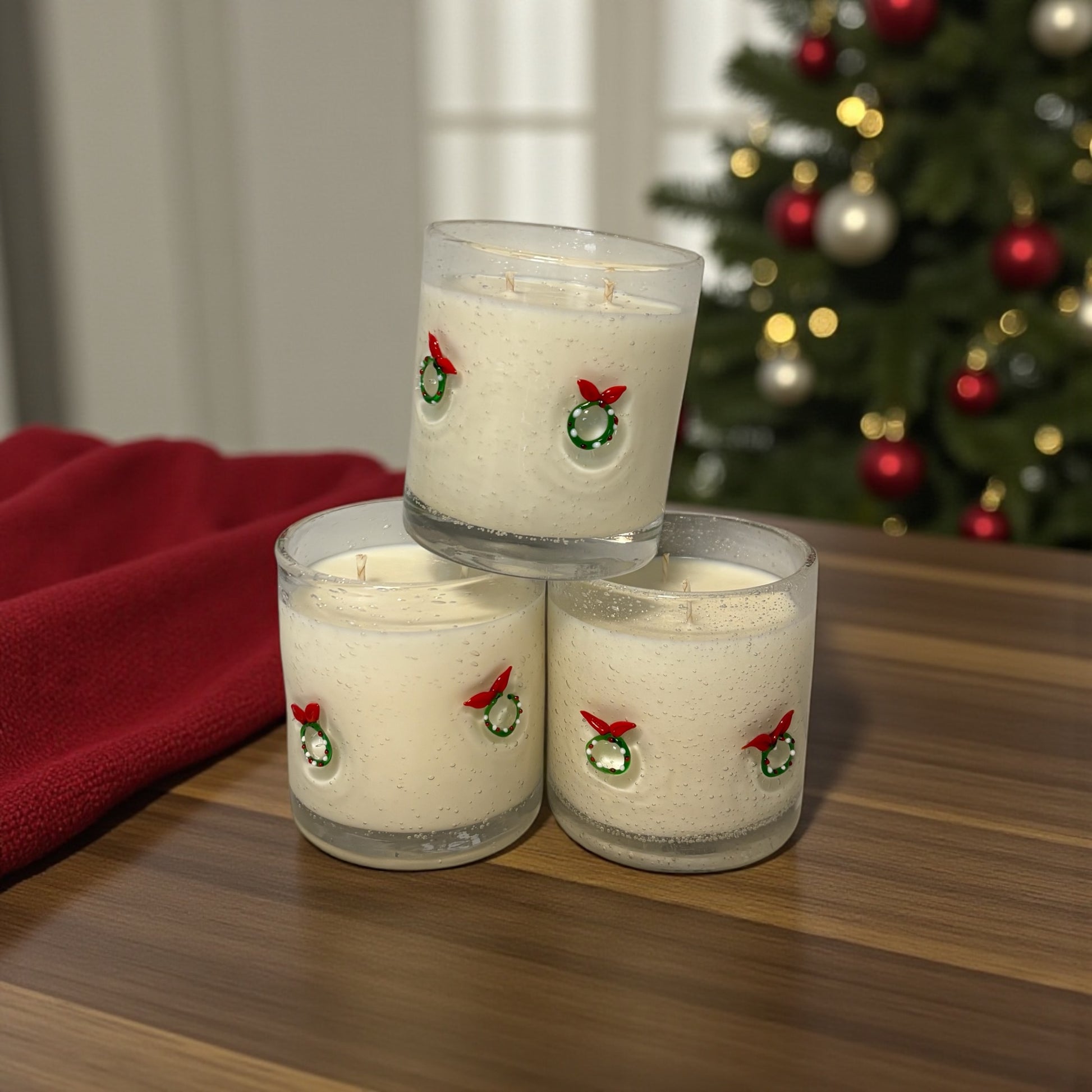 Three candles with Christmas-themed designs stacked on a wooden surface with a decorated tree in the background.