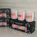 Three hand-poured discovery set candles in pink glass holders with white bases, printed with 'HO!' in holiday colors, displayed on top of their black packaging boxes.