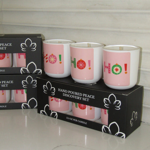 Three hand-poured discovery set candles in pink glass holders with white bases, printed with 'HO!' in holiday colors, displayed on top of their black packaging boxes.