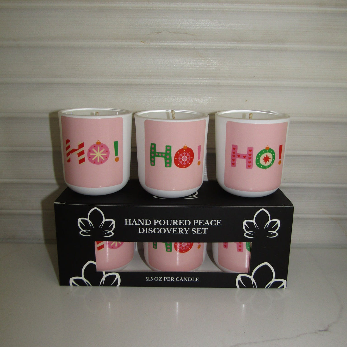 Three pink hand-poured candle jars with 'Ho Ho Ho!' holiday lettering designs, displayed on top of their black retail box labeled 'Hand Poured Peace Discovery Set'.