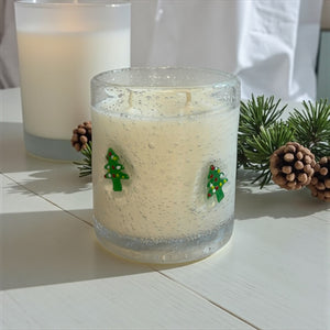 Candle in a glass jar with Christmas tree decorations on a white surface with pine branches and pinecones.