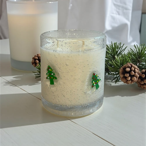 Candle in a glass jar with Christmas tree decorations on a white surface with pine branches and pinecones.