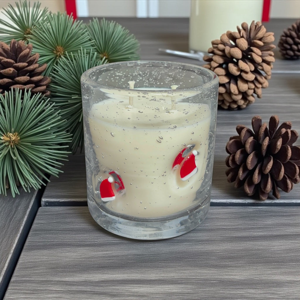 Glass candle with Santa Claus design surrounded by pine cones and greenery on a wooden surface
