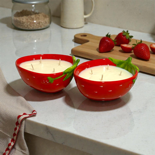 Two red strawberry-shaped candles on a countertop with strawberries and a cutting board in the background.
